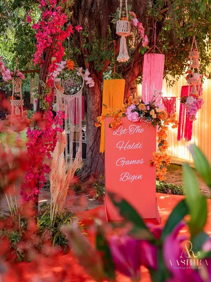 A fun welcome sign for the Haldi ceremony, surrounded by bougainvillea, pampas grass, and hanging macrame decor, setting a playful and festive mood.