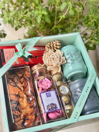 The Scented Bliss hamper is designed for relaxation and indulgence. It includes two festive lanterns, a jar of fragrant potpourri, chocolate-coated almonds, Ferrero Rocher, and a delicious marble tea cake, all tied with a Happy Diwali ribbon.