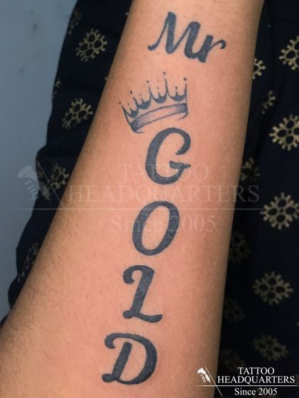 A unique lettering tattoo that spells out "Mr GOLD" vertically down the arm, complete with a crown.
