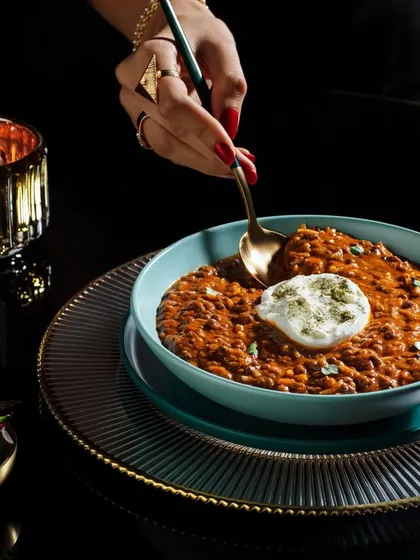 This image of Dal Makhani for Club Jolies uses dark, luxurious tones to create a sense of indulgence. A hand is shown scooping the creamy lentil dish, making it feel warm and inviting.