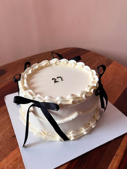 A round cake with a simple and elegant coquette design. The white buttercream is accented with classic piping and black ribbon bows.