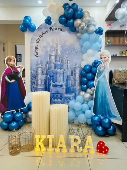 A magical 'Frozen' themed birthday party for Kiara. This setup features cutouts of Anna and Elsa, a castle backdrop, and a balloon arch in icy blue and silver, creating a true winter wonderland.
