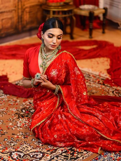 A model seated gracefully on a traditional carpet, surrounded by the flowing fabric of her red saree. This composition highlights the richness of the textures and colors in the frame.