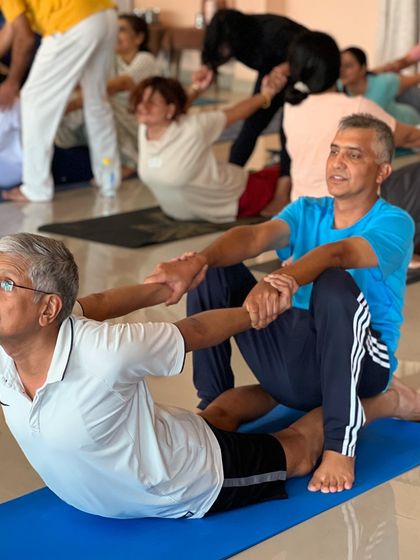 Two students practice a partner version of Dhanurasana (Bow Pose), learning how to support and deepen backbends together.