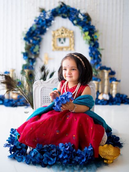 A little Radha dressed in red, sitting gracefully amidst a sea of blue flowers.