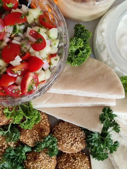 A fresh and vibrant Mezze Platter, perfect for sharing. This spread includes our homemade sesame falafel, soft pita bread, a zesty Israeli salad, creamy hummus, and cool tzatziki dip.