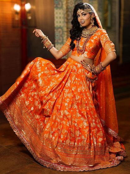 A full-length portrait showcasing a vibrant orange Banarasi lehenga. The makeup features a dramatic smokey eye and bold jewelry, creating a stunning look for a Sangeet or wedding ceremony.