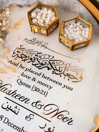 An elegant white and gold ring tray with delicate daisy-like flowers and a beautiful Quranic verse. The hexagonal ring boxes are filled with pearls.