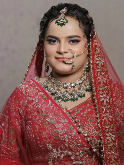 A close-up portrait of a beautiful bride. Her makeup features defined eyes and a soft smile, perfectly complementing her red lehenga and green-beaded jewelry.