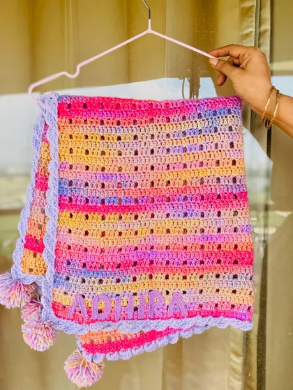 The "Adhira" blanket hanging, showcasing the vibrant colors and the soft, cuddly texture of the yarn. This custom piece is a wonderful example of a personalized baby shower gift.