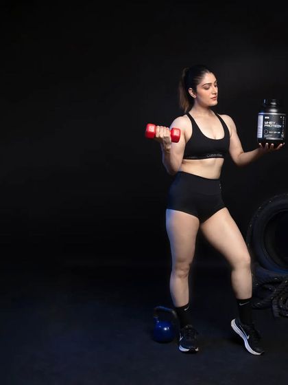 This image combines action with product focus. The model performs a bicep curl with a dumbbell while holding the whey protein in her other hand, visually connecting the product directly to the act of muscle building.