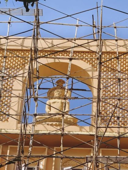 A close-up view of the Jain Upashray's facade during construction. This shot highlights the detailed craftsmanship of the stone statue and the surrounding jali work, framed by the scaffolding that supports its creation.