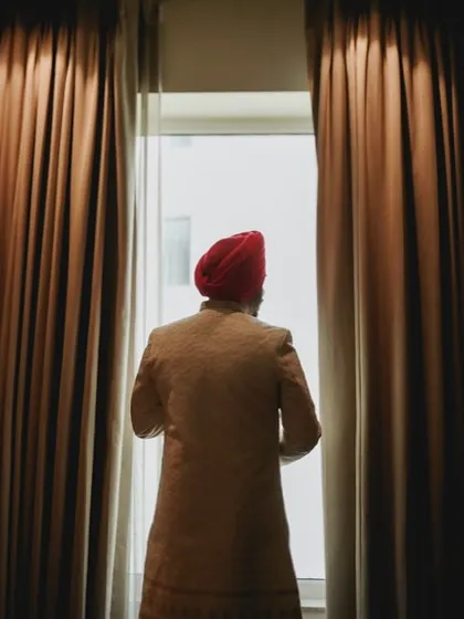 The groom looks out the window, a contemplative moment before the wedding ceremony begins. The curtains frame him perfectly.