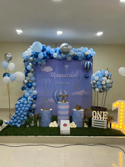 A "Hot Air Balloon" themed first birthday decoration. The setup features a custom printed backdrop, a balloon garland in shades of blue and silver, and a creative hot air balloon structure made of balloons and a crate.