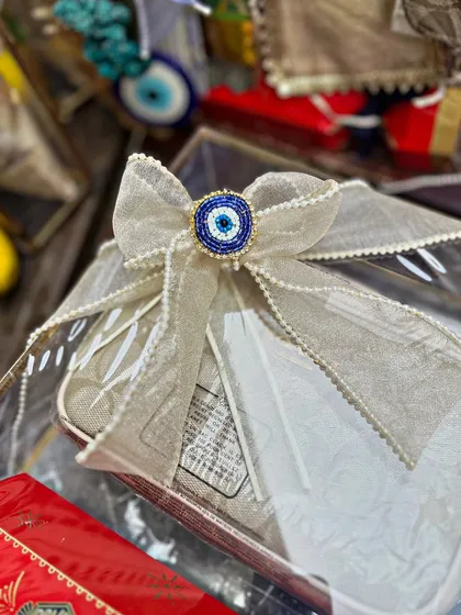 A close-up of a beautiful bow made from textured ribbon and centered with a beaded evil eye charm. This detail, placed on a transparent gift box, adds a touch of bespoke luxury.