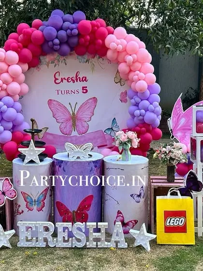 An outdoor butterfly-themed party with a beautiful balloon arch in shades of pink and purple. The setup includes a small white picket fence and butterfly props for a garden feel.