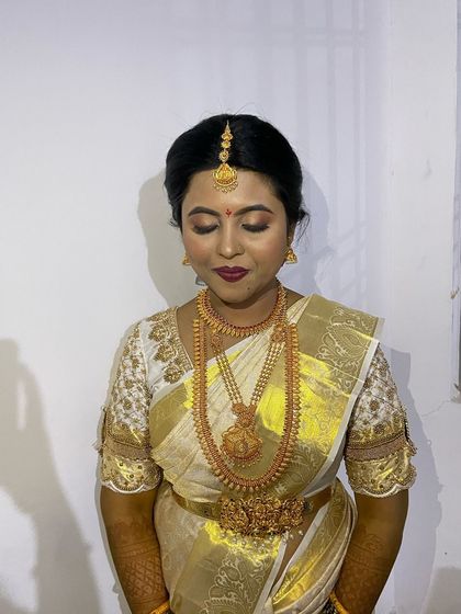 A beautiful muhurtham look for Vedavathy. The makeup is classic and clean, with a deep plum lip and defined eyes that complement her off-white and gold saree.