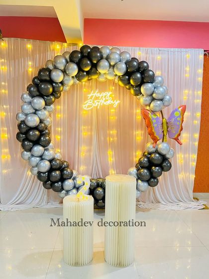 This simple yet elegant birthday backdrop uses a circular frame with black and silver balloons. A large butterfly foil balloon adds a whimsical touch to the design, which is set against a white curtain with fairy lights.
