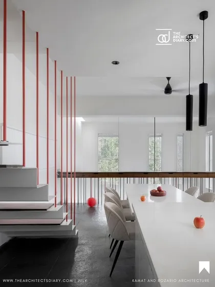 The minimalist dining area and staircase are defined by clean lines and a simple color palette. The vertical red poles of the stair railing create a graphic element that is both functional and artistic, adding a dynamic touch to the space.