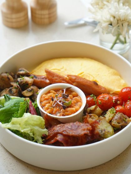 Our breakfast bowl is a complete meal, featuring a classic omelette, sausages, bacon, sautéed mushrooms, baked beans, and fresh greens. It’s the perfect fuel for a busy day.