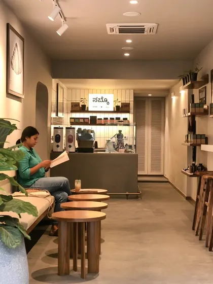 The interior of the Bombay Shirt Company and Subko Coffee collaboration in Indiranagar. The space is designed as a calm, neutral canvas with warm wood furniture and soft lighting, creating a relaxed atmosphere for both shopping and coffee.