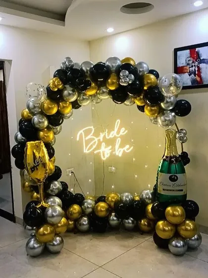 A glamorous bachelorette party backdrop featuring a balloon ring in a striking black, gold, and silver color scheme. The setup is accented with champagne and wine glass foil balloons for a fun, celebratory theme.