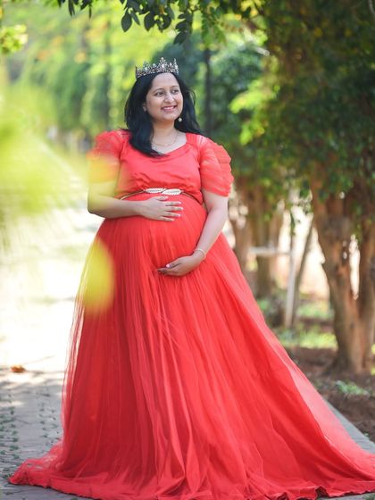 A joyful portrait of a mom-to-be in a red gown and crown, smiling brightly in a park.