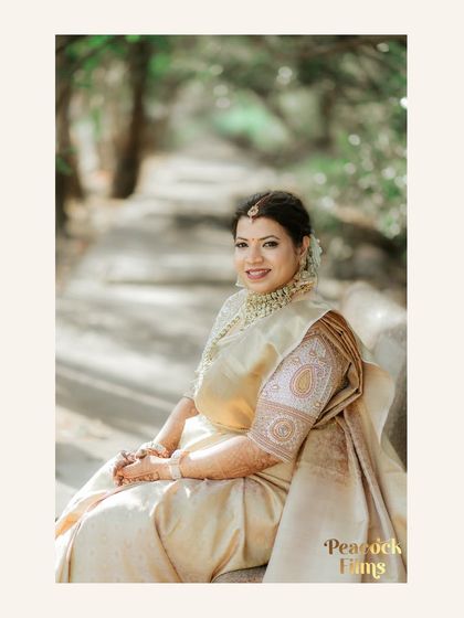 A serene portrait of the bride in her elegant off-white and gold saree, seated in a peaceful outdoor setting.