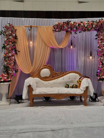 A modern and chic engagement stage design. The combination of peach draping, an asymmetrical floral arch, and industrial-style hanging lights creates a sophisticated and stylish backdrop.