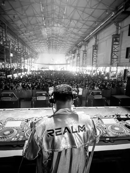 A shot from behind the decks at a massive warehouse-style festival. The raw, industrial setting combined with a huge crowd creates an intense and immersive techno experience.