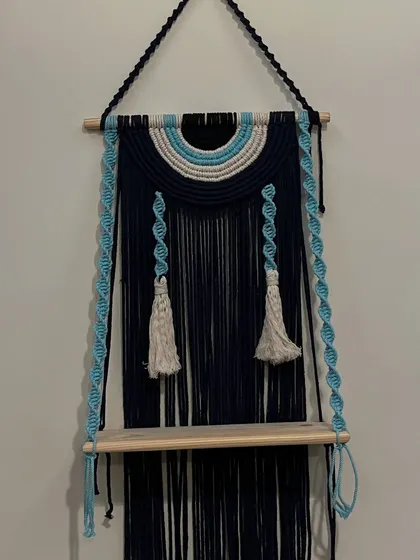 I love creating pieces that are both beautiful and useful. This evil eye themed wall shelf does just that, combining a protective symbol with a practical wooden shelf. The deep navy blue and sky blue cords with twisted hangers make it a unique piece to display small plants or treasures.