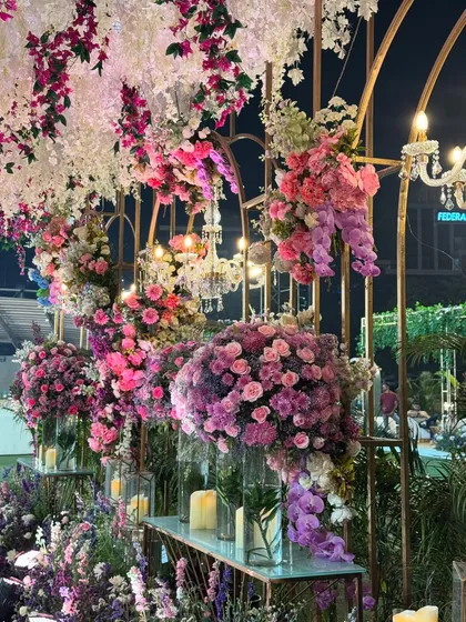 A beautiful installation of pink, purple, and white flowers cascading over a golden archway. The mix of colors and textures creates a rich, romantic, and whimsical feel. 🩷💜