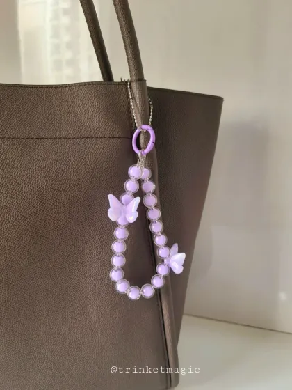 A lovely lavender-colored beaded bag charm featuring two purple butterfly accents. It's shown on a tote bag, adding a touch of pastel magic.