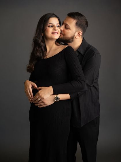 An intimate studio portrait with the couple dressed in classic black. His gentle kiss on her shoulder and their hands cradling the bump make for a timeless and romantic image.