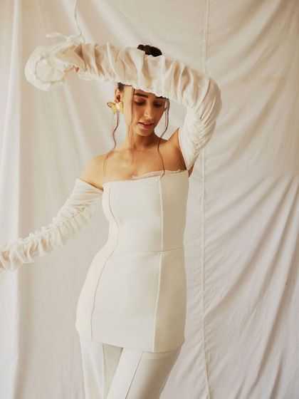 An all-white look from a creative fashion shoot. The styling, featuring a structured top with dramatic, ruched off-shoulder sleeves, creates an ethereal and sculptural silhouette against the simple white backdrop.