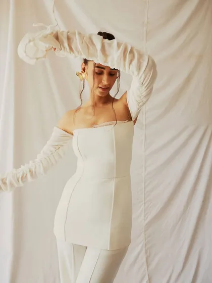An all-white look from a creative fashion shoot. The styling, featuring a structured top with dramatic, ruched off-shoulder sleeves, creates an ethereal and sculptural silhouette against the simple white backdrop.