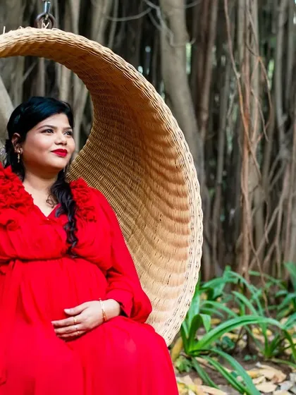 A serene portrait of the mom-to-be in a vibrant red dress, seated in a hanging wicker chair, surrounded by the unique texture of banyan tree roots.