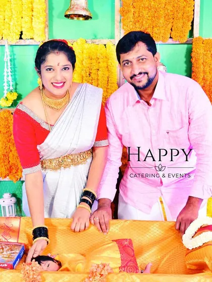 A happy couple during their baby's naming ceremony. They stand behind the cradle against a traditional backdrop of marigold flower panels.