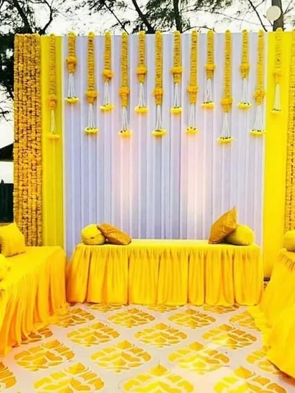 A bright and sunny Haldi ceremony setup with a yellow and white color scheme. The backdrop features hanging floral strings made of marigolds, creating a traditional and festive atmosphere.