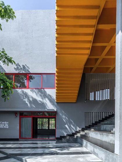 L'Atelier at Greenwood High School uses a simple palette of grey plaster punctuated by bold primary colors. The yellow underside of the staircase creates a vibrant, playful intervention, reflecting the creative energy of the art and music school within.
