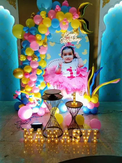A colorful and personalized first birthday decoration at home. This setup features a custom backdrop with the baby's photo, a vibrant balloon arch, and light-up marquee letters.