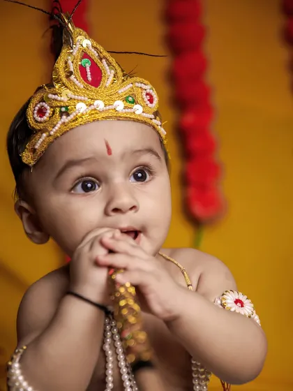 A beautiful close-up of a baby Krishna with wide, curious eyes. The soft lighting and warm yellow background highlight his innocent features.