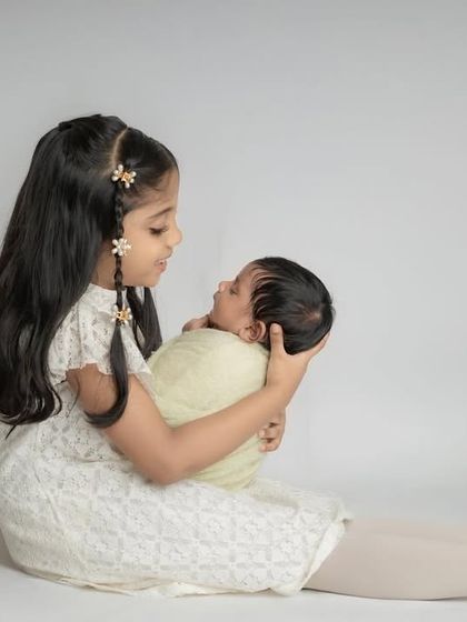An older sister sits on the floor and holds her newborn sibling, looking down at them with a gentle and loving expression.