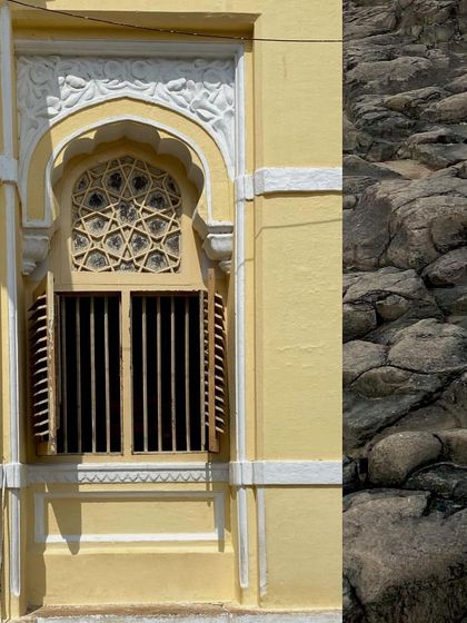An ornate window on a yellow heritage building, next to a textured rock wall.