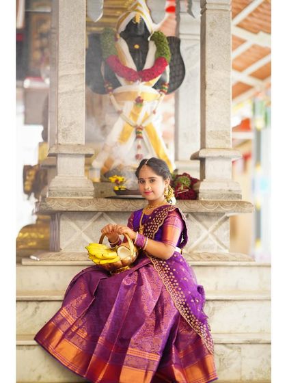 A young girl in a traditional purple saree sits on the steps of a temple, holding a basket of offerings for a cultural photoshoot.