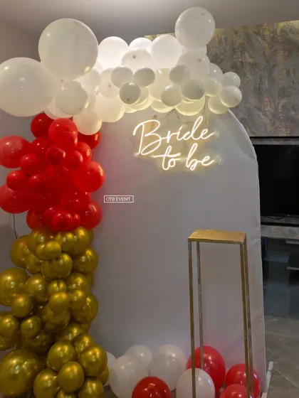 A bold and classic 'Bride to be' backdrop featuring a red, white, and gold balloon garland. The neon sign on the white arched panel really pops.