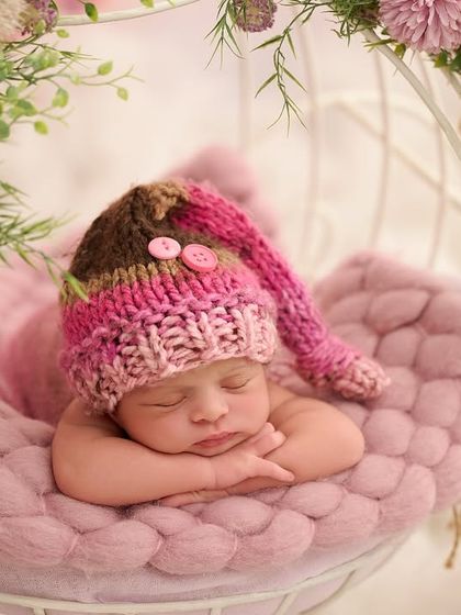 This close-up shot features a baby in a pink and brown knitted hat, sleeping in a white wire basket decorated with pink flowers and soft textures.
