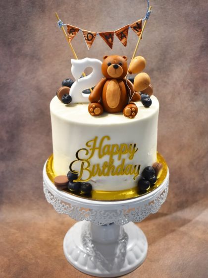 An adorable teddy bear-themed cake for a 2nd birthday. The cake is topped with a cute fondant teddy bear holding balloons, a mini bunting, and the number 2.