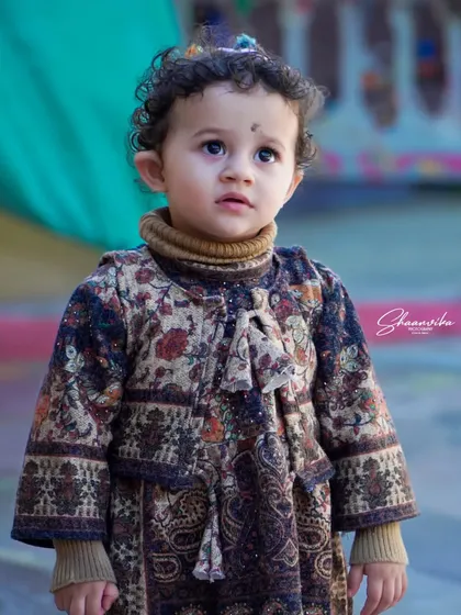 A close-up portrait of a toddler looking up with wide, curious eyes. The soft lighting highlights her delicate features and traditional-style clothing.