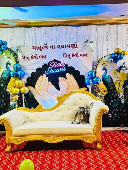 An elegant and traditional baby shower setup. This design features a beautiful peacock theme with blue, green, and gold balloon arrangements, a custom backdrop with Gujarati blessings, and a classic chaise lounge for the mother-to-be.
